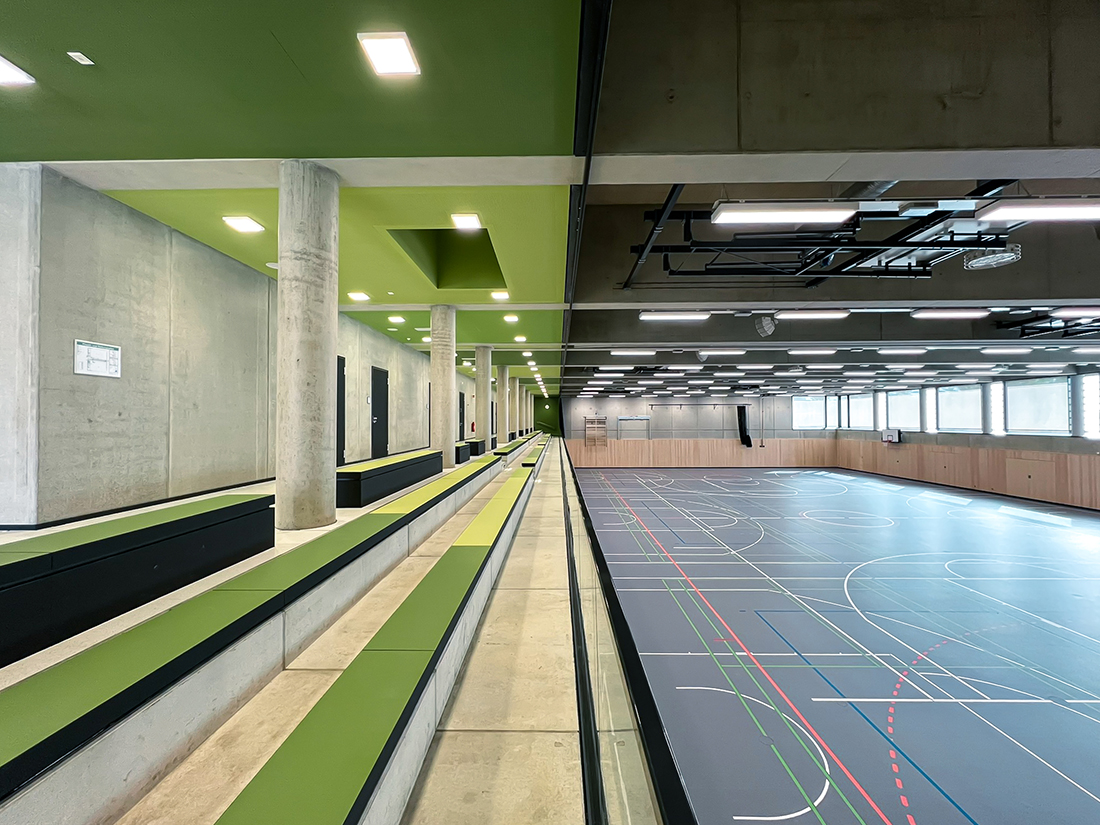 Innenansicht einer modernen Dreifachsporthalle mit breiter Tribüne auf der linken Seite. Die Halle ist durch klare Linien, Sichtbeton, grüne Farbakzente an Decke und Sitzflächen sowie durch großzügige Beleuchtung und Fenster geprägt. Das Spielfeld zeigt verschiedene Linienmarkierungen für mehrere Sportarten.