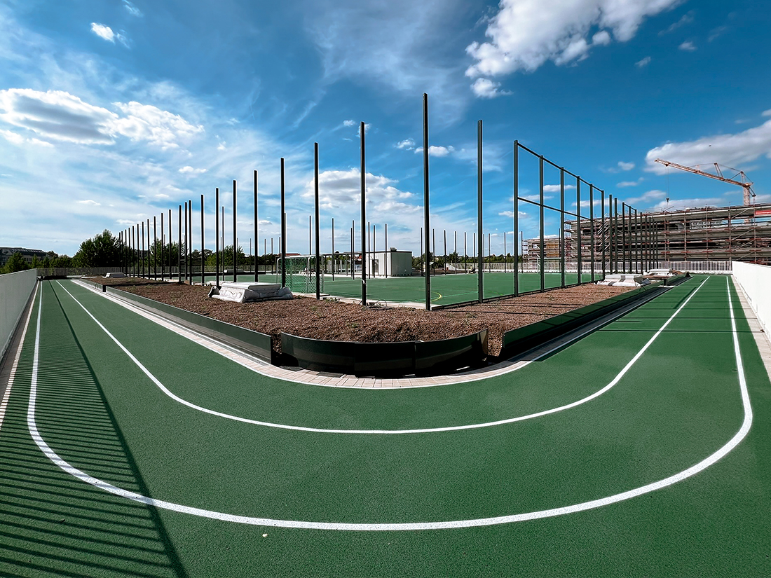 Zu sehen ist eine Sportanlage auf einem begrünten Dach mit einer umlaufenden Laufbahn mit grünem Belag und weißen Markierungen. Zwischen der Laufbahn und dem Fußball- bzw. Basketballfeld befinden sich Pflanzflächen sowie Pfosten, die für Ballfangnetze oder Begrenzungen vorgesehen sind. Im Hintergrund erkennt man weitere Sportflächen und Bäume. Der Himmel ist blau mit einigen weißen Wolken.